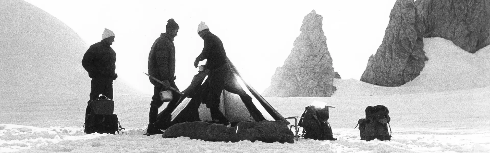 Drie mensen in winterkleding staan bij een tent en rugzakken in een besneeuwd landschap, met grote rotsen op de achtergrond. Het tafereel straalt kou en afzondering uit, en de figuren steken als silho